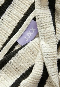 Pletený sveter s horizontálnymi pruhmi v krémovej a čiernej farbe. Má textúrovanú látku a levanduľový štítok s nápisom "JUXX" v bielej farbe.