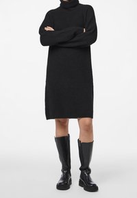 Robe pull noire à col roulé avec une coupe décontractée, longueur au genou et texture côtelée. Portée avec des bottes noires montant jusqu'aux genoux.