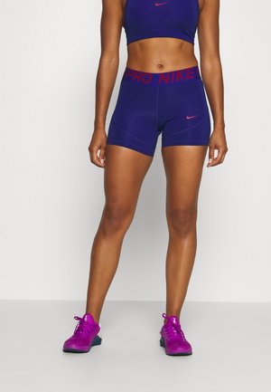 Vrouw draagt paarse Nike sportshorts, een cropped top en felpaarse sportschoenen, staand op een effen achtergrond.