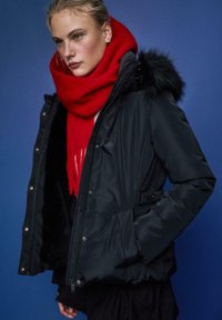 Črna puhasta jakna s kapuco obloženo z umetnim krznom, z dvema sprednjima žepoma in sijočim zaključkom, kombinirana s svetlo rdečim volnenim šalom. Modro ozadje.