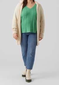 Cremfarbener Strickcardigan, grünes geripptes V-Ausschnitt-Top, blaue Jeans und beigefarbene Ankelboots mit schwarzer Sohle. Strukturierte Materialien und gestuftes Design.