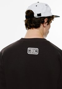 Sort sweatshirt med rund halsudskæring og ribbede manchetter. Har et hvidt broderet patch på ryggen med teksten "RADICAL NEW VISIONS."