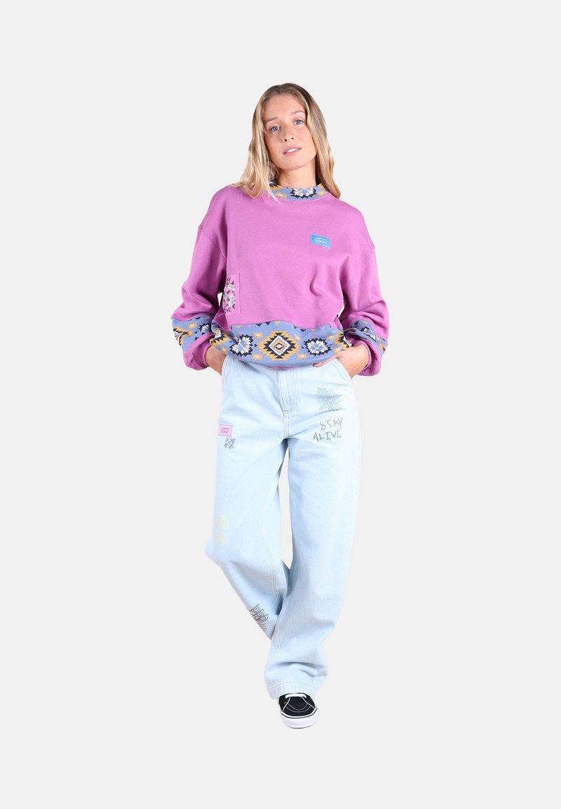 Sweat-shirt violet avec des poignets et un ourlet à motifs, associé à un jean baggy bleu clair avec des détails brodés, et des chaussures noires.
