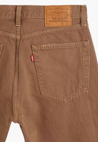 Pantalones de denim de algodón marrón en tejido twill con una etiqueta de cuero en la cintura, que cuentan con dos bolsillos traseros con un patrón de costura distintivo y una etiqueta roja.