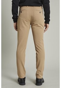 Slim-fit, beżowe spodnie o gładkiej teksturze. Posiadają podwójne tylne kieszenie z czarnymi guzikami oraz subtelnymi detalami szycia.