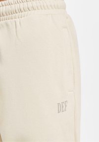 Lyse beige sweatpants med elastisk talje, der har et diskret "DEF" logo i grå, samt en sidelomme. Blød tekstur.
