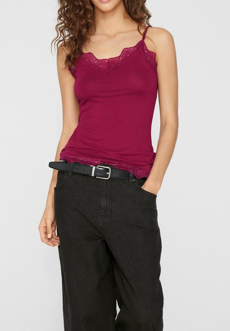 Femme portant un caraco ajusté rose foncé bordé de dentelle, rentré dans un pantalon noir ample taille haute avec une ceinture noire.