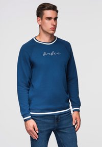 Muž v modrém mikině s bílými pruhy na manžetách a dolním lemu, označené "Ombre," v kombinaci s modrými džíny, stojící na jednobarevném pozadí.