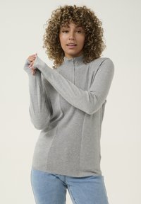 Pilkas zip-up megztinis su braižytos tekstūros ir ilgomis rankovėmis. Turinti aukštą apykaklę ir patogų stovą, derinama su šviesiai mėlynomis džinsomis.