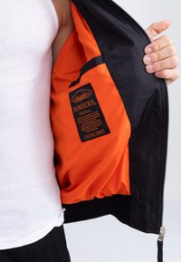 Schwarze Bomberjacke mit orangefarbenem Futter, ausgestattet mit einem Etikett, das "BOMBERS" liest, und einer seitlichen Tasche. Reißverschlussdetails auf der Vorderseite.