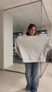 Woman standing indoors holding a large gray sweater stretched wide in front of her, with glass walls and tiled ceiling behind.