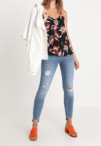 Frau trägt zerrissene blaue Skinny Jeans, schwarzes florales Trägertop, hält eine weiße Jacke über der Schulter und schwarze offene Schuhe mit orangen Absätzen.