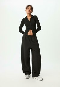 Femme se tenant debout avec les mains derrière le dos, portant une veste noire zippée courte, un pantalon noir ample et des baskets argentées, sur un fond blanc uni.