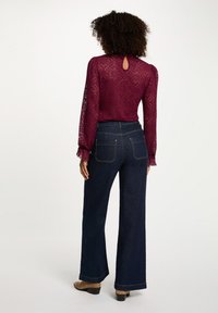 Blusa in pizzo bordeaux con maniche lunghe e un'apertura a chiave sulla schiena, abbinata a jeans a gamba ampia di un blu scuro con tasche posteriori e stivaletti beige.