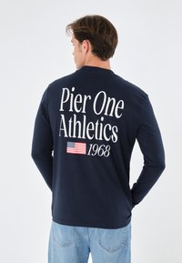 Marineblaues Langarmshirt mit weißem Text "Pier One Athletics 1968" und Grafik der amerikanischen Flagge auf der Rückseite. Weiches Baumwollmaterial.