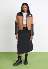 Barna és fekete pufferszövet dzseki cipzárral, bordás mandzsettákkal, szürke crop top, fekete maxi szoknya, és fekete fűzős katonai csizma zöld padlón.