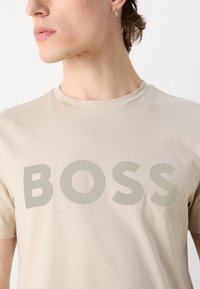 Beige színű, pamut póló, amelyen egy nagy, szürke "BOSS" felirat található elöl. Klasszikus kerek nyakkivágással és rövid ujjal rendelkezik. Sima textúrájú.