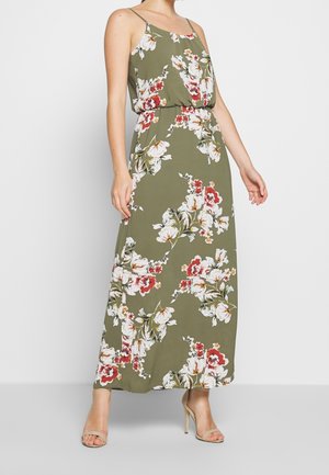 Femme portant une robe longue olive à imprimé floral blanc et rouge avec de fines bretelles, associée à des sandales à talons hauts beiges.