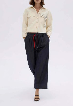 Donna con camicia corta color crema con bottoni e dettaglio tasca, pantaloni neri a gamba larga con coulisse rossa, sandali neri con cinturini.