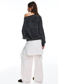 Kobieta w luźnym, ciemnym, puszystym swetrze zsuwającym się z jednego ramienia, białej warstwowej spódnicy i szerokich kremowych spodniach, stojąca na białym tle.