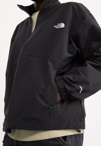 Chaqueta impermeable negra con cierre de cremallera, puños elásticos y dos bolsillos laterales. Presenta un logotipo blanco en el área izquierda del pecho.