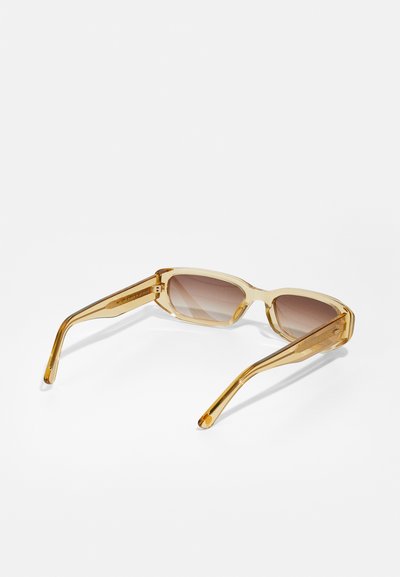 Ochelari de soare translucizi galbeni cu lentile maro în gradient. Design unghiular cu brațe drepte, având un finisaj lucios și un detaliu subtil al logo-ului.