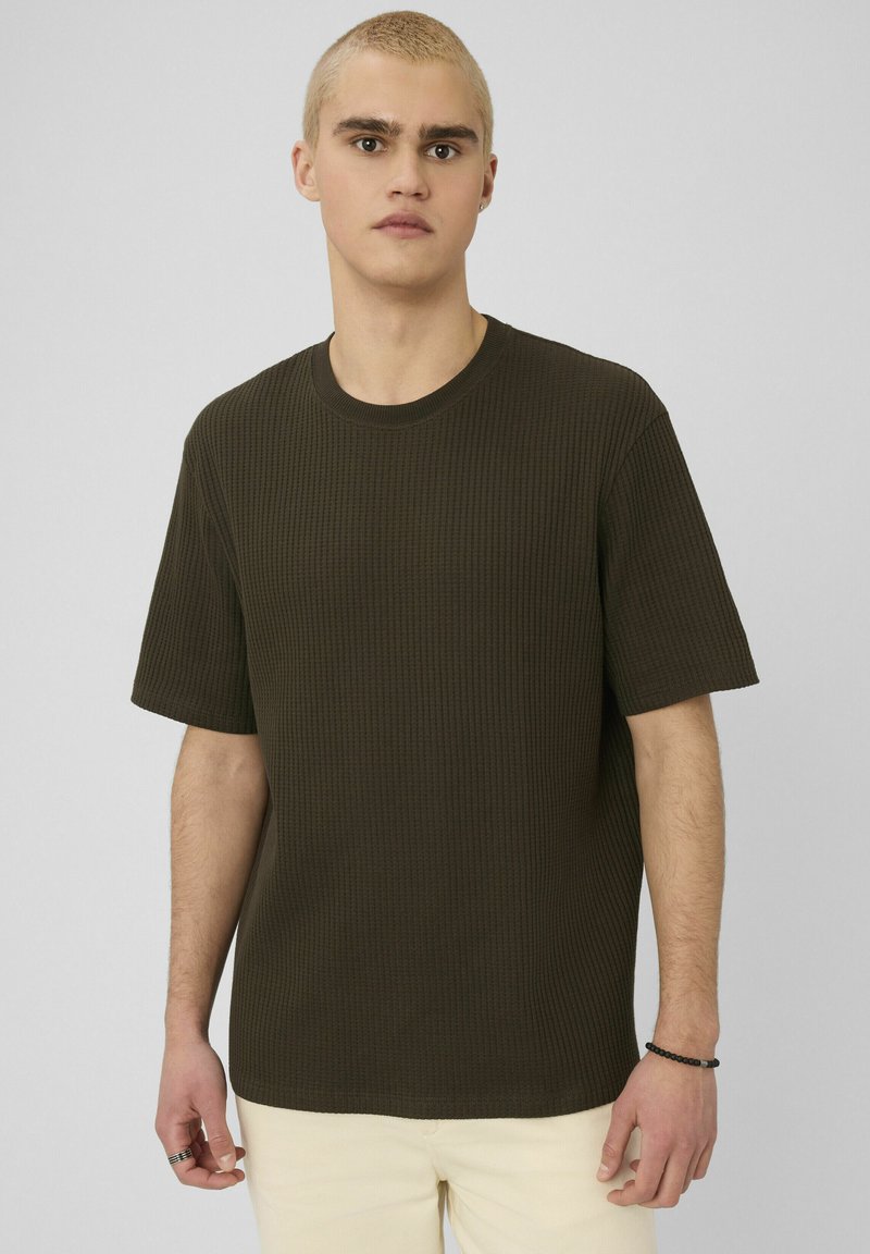 Jeune homme aux cheveux blonds courts portant une chemise texturée à manches courtes de couleur vert foncé et un pantalon de couleur claire, sur un fond uni.