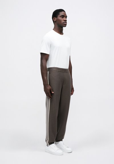 Άνδρας που στέκεται στραμμένος προς τα δεξιά, φορεμένος με λευκό μπλουζάκι, καφέ αθλητικά παντελόνια με πλευρικές ρίγες και λευκά αθλητικά παπούτσια, σε απλό λευκό φόντο.