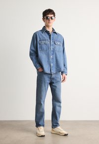Chemise en denim avec un devant boutonné, deux poches poitrine ; assortie à un jean bleu et des baskets crème avec des semelles foncées. Le cadre a une texture légère.