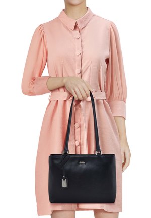 Woman in a pink buttoned dress holding a black leather handbag with two handles and a hanging tag in front of her.