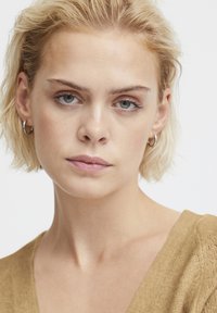 Nahaufnahme einer Person mit kurzen, welligen blonden Haaren, die ein beiges Strickoberteil und silberne kreisförmige Ohrringe trägt. Sie hat klare blaue Augen und ein natürliches Make-up.