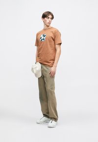 Καφέ βαμβακερό T-shirt με γραφικό λογότυπο, συνδυασμένο με ανοιχτό μπεζ παντελόνι. Ένα κρεμ καπέλο κρατιέται στο ένα χέρι. Λευκά αθλητικά παπούτσια ολοκληρώνουν την εμφάνιση.