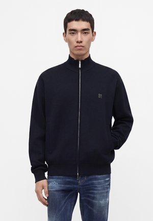 Junger Mann trägt eine schwarze Zip-Jacke mit kleinem Logo auf der Brust und blaue verwaschene Jeans, steht mit einer Hand in der Tasche.