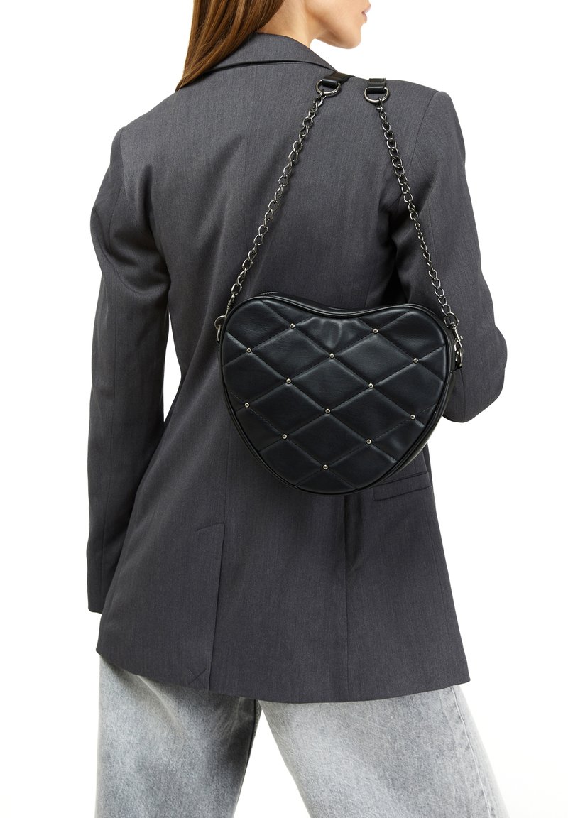 Borsa a forma di cuore nera realizzata in pelle trapuntata con borchie argentate, dotata di una tracolla a catena e finitura lucida. Indossata sopra un outfit con blazer grigio.