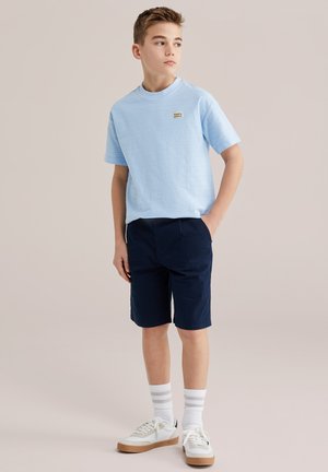 Niño con camiseta azul claro con el logo "Puerto Montana", pantalones cortos azul marino, calcetines blancos con rayas y zapatillas blancas, de pie con una mano en el bolsillo.