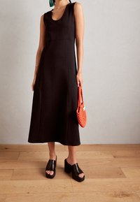 Musta ilmanhiha dressi, jossa on pyöreä pääntie, A-linjainen muoto ja sileä pinta. Mallilla on käsissään pieni oranssi kudottu käsilaukku ja jalassaan mustat korkokengät.