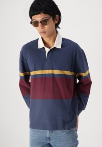 Langermet polo skjorte i marineblå med hvit krage, med horisontale striper i maroon, mørkeblå og sennepsgul. Laget av bomull.