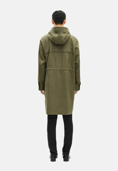 Uomo che indossa un lungo giubbotto con cappuccio verde oliva, pantaloni neri e scarpe nere, in piedi di spalle su uno sfondo bianco puro.