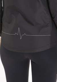 Sort atletisk jakke med en hjerterytmelinje i hvid langs kanten. Stoffet er glat med en tætsiddende pasform. Sorte leggings er synlige.