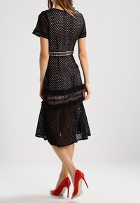 Kvinna som står med ryggen synlig, iförd en svart perforerad midiklänning med spetsdetaljer och röda högklackade skor.