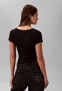 Pruunide juustega naine kannab musta lühikese varrukaga t-särki ja musti teksaseid, millel on taskul tikitud CK logo, seistes halli tausta ees.