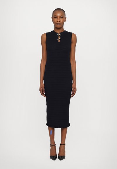Robe en tricot côtelé marine avec un col rond et un détail en forme de clé, dotée d'un ourlet froncé et associée à des escarpins noirs à bout pointu.