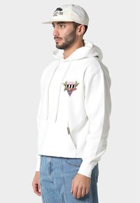 Biała bluza z kapturem z grafiką na przedzie, przedstawiającą paski, liście i trójkąt. Posiada sznurek w kapturze oraz przednią kieszeń. Noszona z jasnoniebieskimi dżinsami.