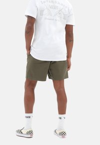 Hvid t-shirt med et trykt design på ryggen, olivengrønne shorts og ternede slip-on sko. Synlige sokker med logo.