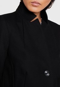 Blazer noir avec col montant, texture côtelée et une fermeture à un bouton noir. Vue en gros plan mettant en évidence le tissu et le design du col.