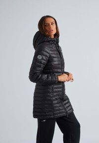 Zwarte gewatteerde winterjas met een capuchon. Heeft een gestikte structuur en een ritssluiting. Inclusief een embleem op de bovenste mouw.