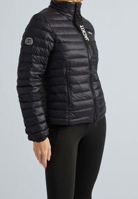 Femme portant une veste matelassée noire avec des poches zippées et des logos sur la poitrine et le haut du bras gauche.