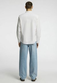 Balta mygtukų marškiniai su tekstūruotu paviršiumi ir ilgomis rankovėmis, derinami su šviesiai mėlynomis plačiomis džinsomis ir juodais batais, matomi iš nugaros.