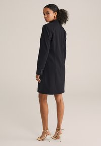 Rochie bleumarin tip sacou cu un design ajustat, mâneci lungi și manșete cu nasturi. Material neted cu o siluetă clasică. Tocuri aurii completează ținuta.