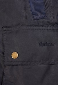 Černá kapsa na bundě Barbour se kovovým patentním knoflíkem s vyraženým nápisem "Barbour" a černým vyšitým logem "Barbour" na látce.